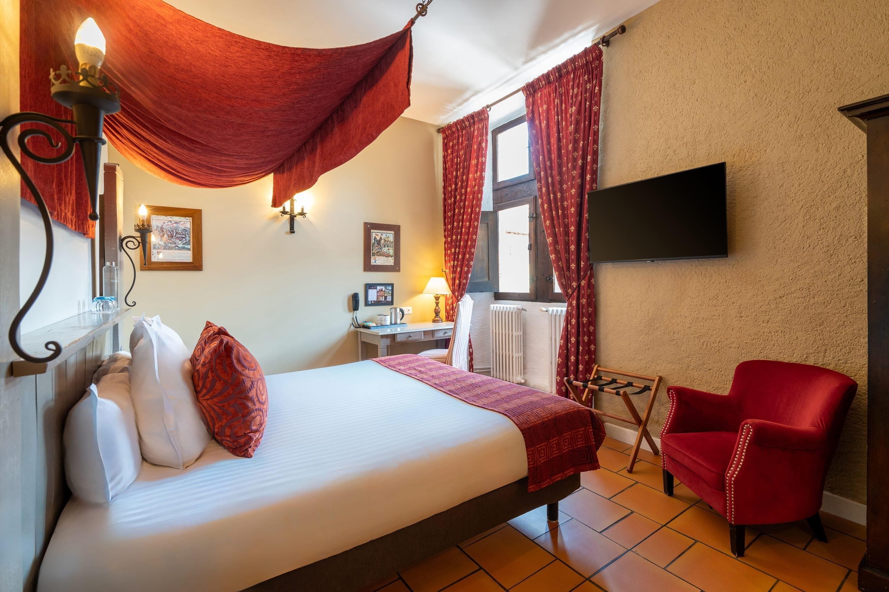 Habitación de hotel con una cama, una silla roja, un escritorio con lámpara y un televisor montado en la pared.