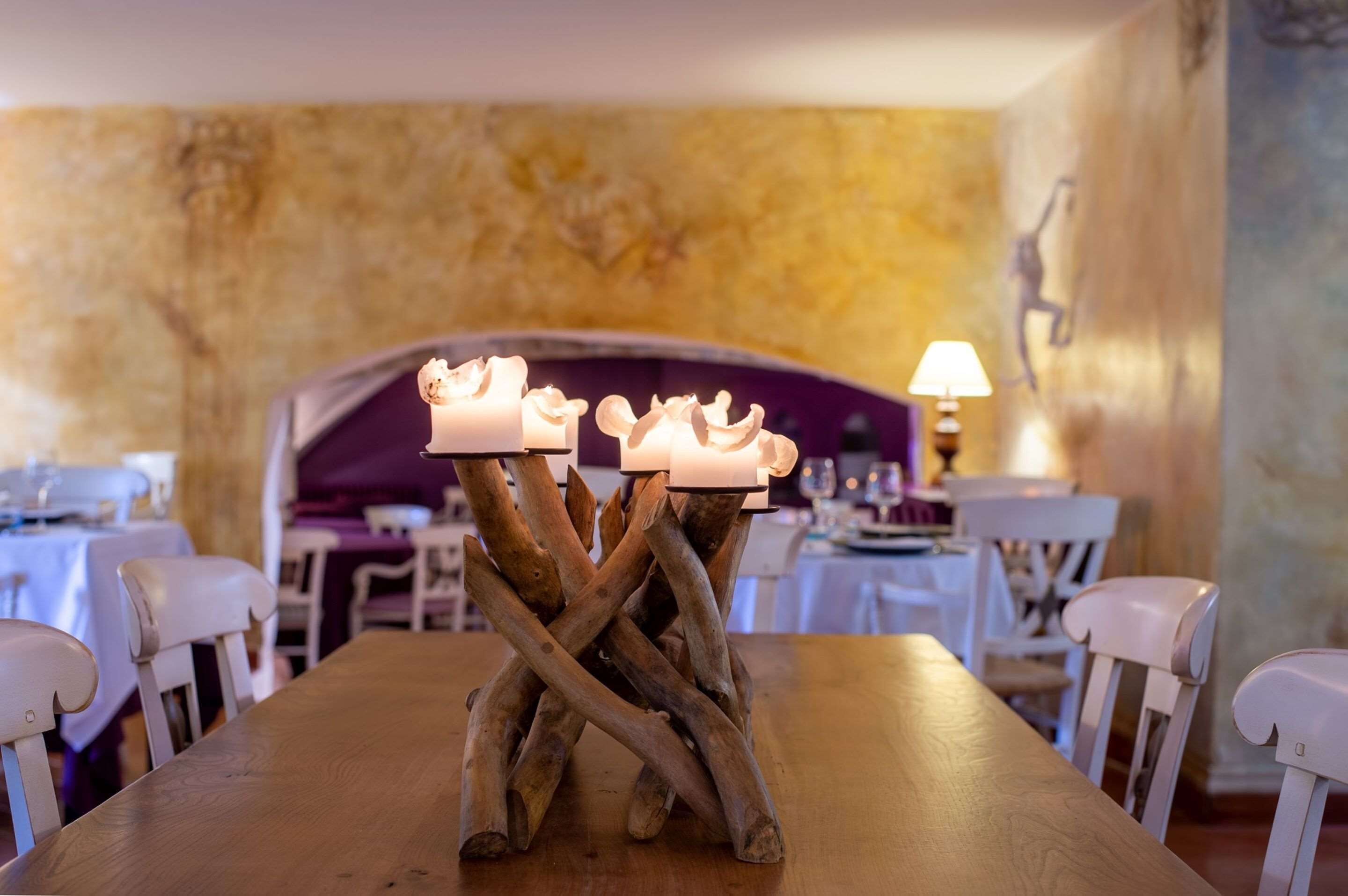 Una mesa de comedor con un llamativo centro de ramas entrelazadas sosteniendo velas, rodeada de sillas blancas en una habitación con pared texturizada y un sofá púrpura al fondo.