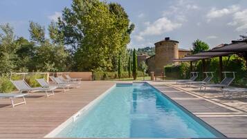 Une piscine rectangulaire bordée de chaises longues sur des terrasses en bois, entourée de verdure et avec un bâtiment en pierre en arrière-plan.