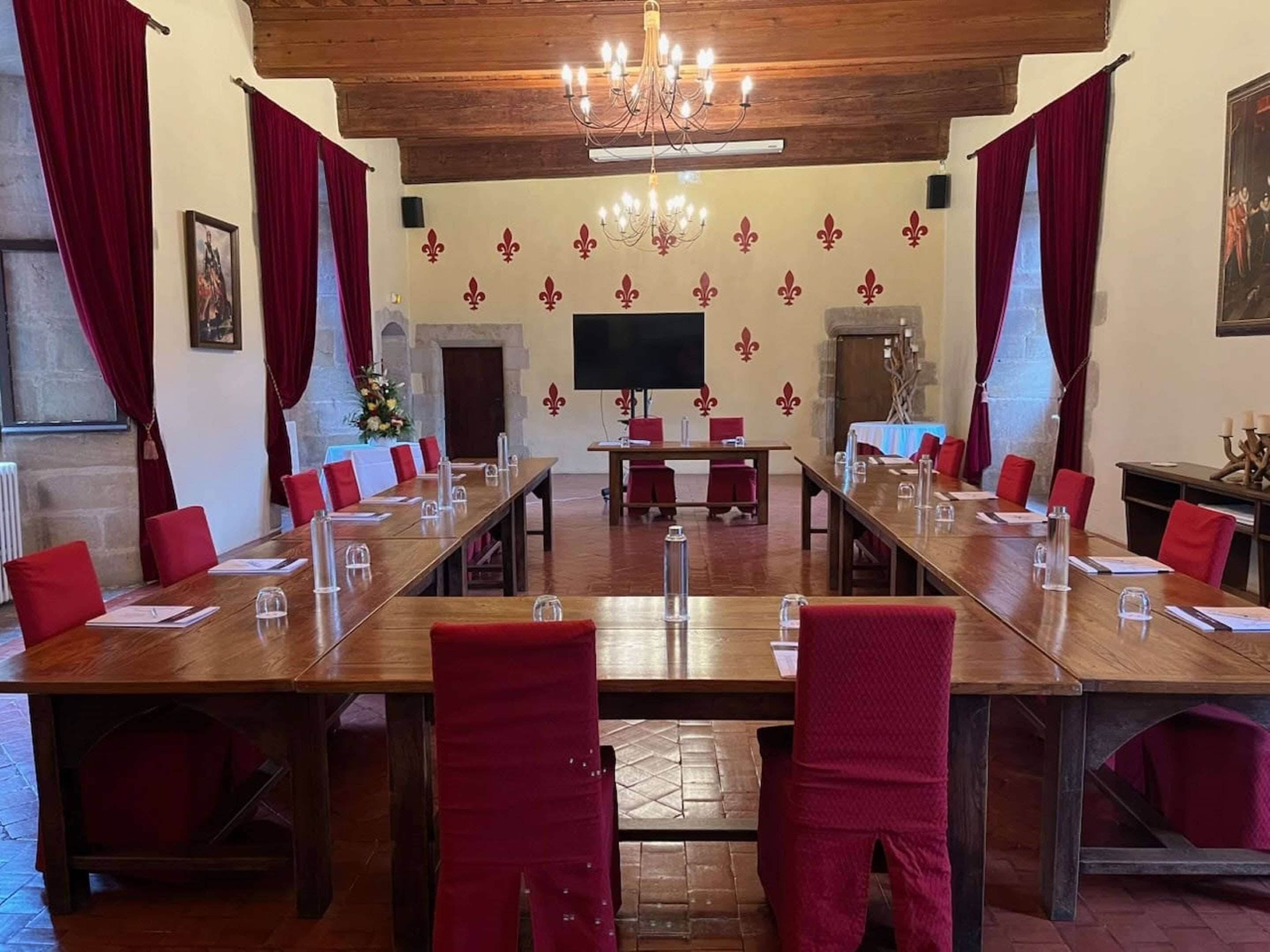 Une salle de conférence avec de longues tables, des chaises rouges, un lustre et un grand écran.