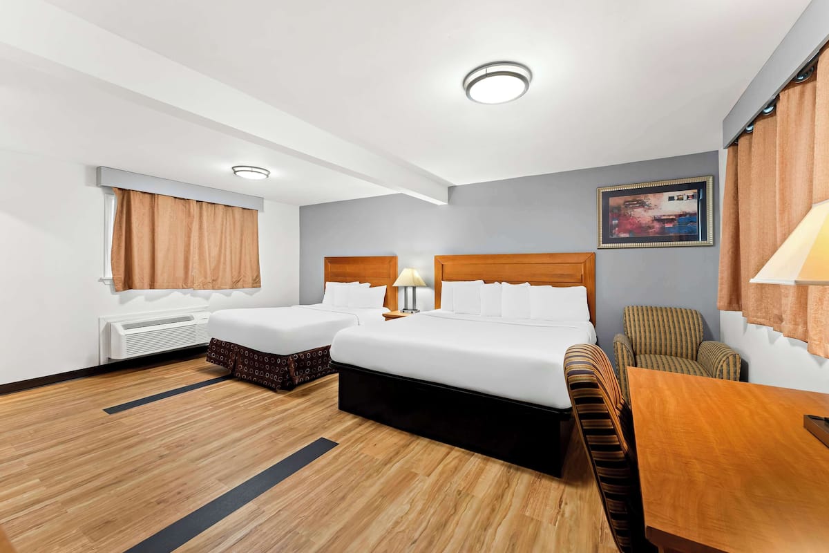 A hotel room with two beds, a wooden floor, a desk, a chair, a lamp, a framed picture on the wall, and a window with curtains.