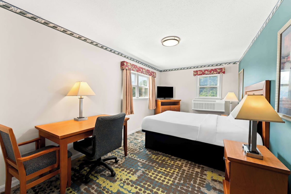 A hotel room with a bed, desk, chair, two lamps, an air conditioning unit, and a window with curtains.