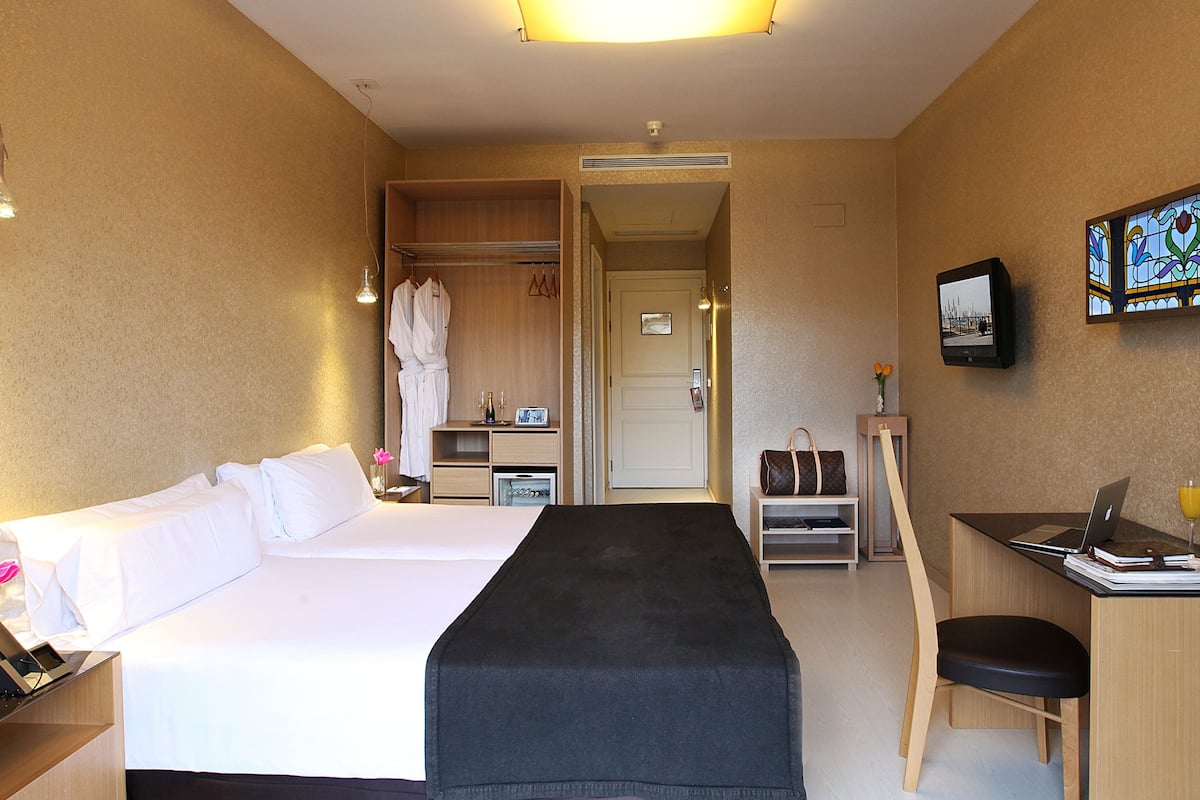 A hotel room with a bed, a desk with a laptop, a chair, a robe, and a coat rack.