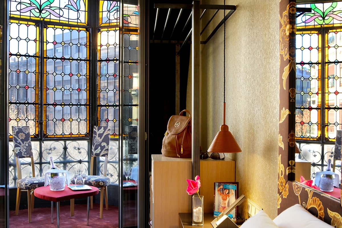 A hotel room with a large window featuring stained glass, a wooden nightstand with a lamp, a chair, and a small table with a vase of flowers.