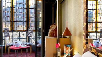 Habitación de hotel con un ventanal grande con vidrieras, una mesita de noche de madera con una lámpara, una silla y una pequeña mesa con un jarrón de flores.