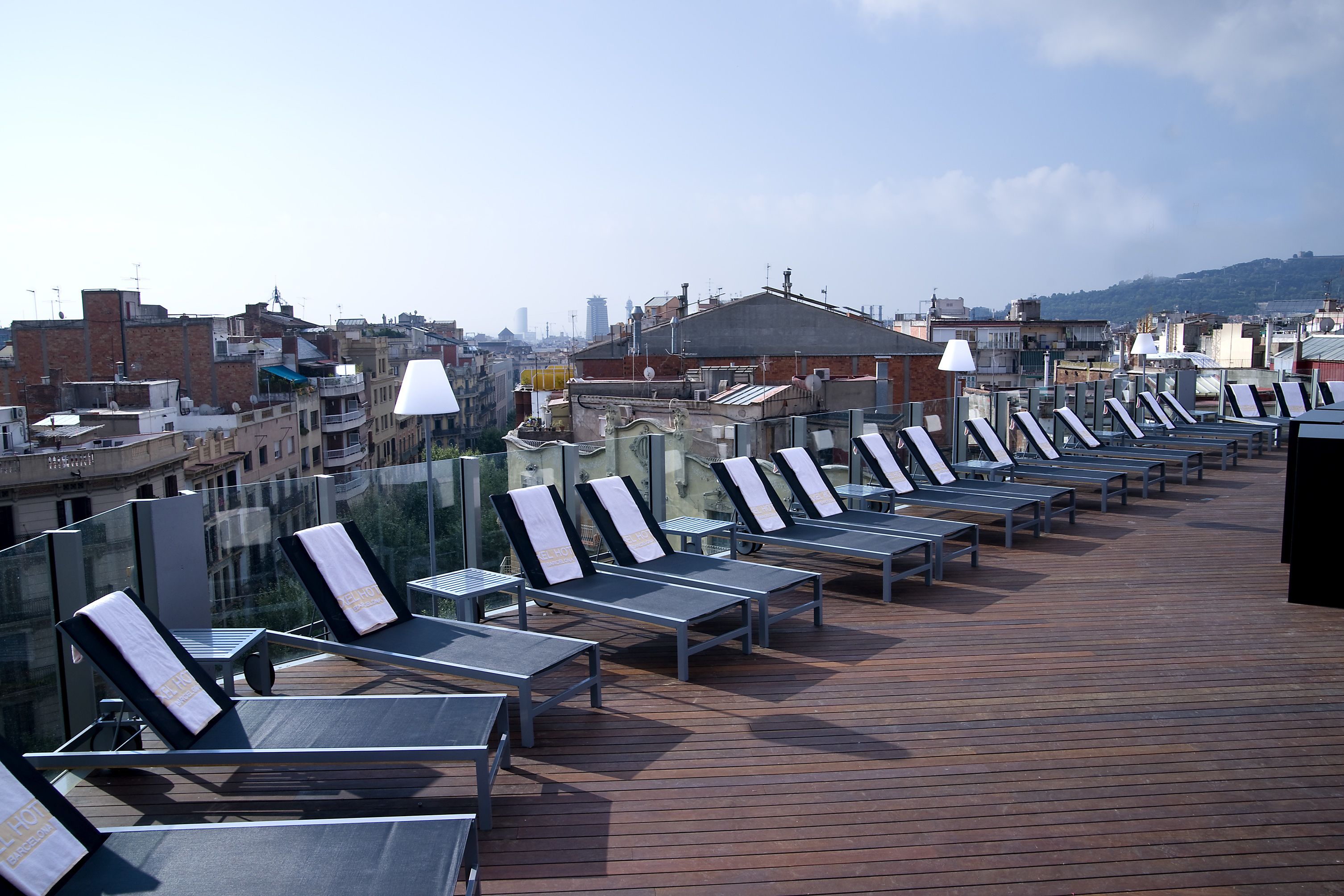 A rooftop terrace with sun loungers and a wooden deck, overlooking a cityscape with buildings and a distant hill.