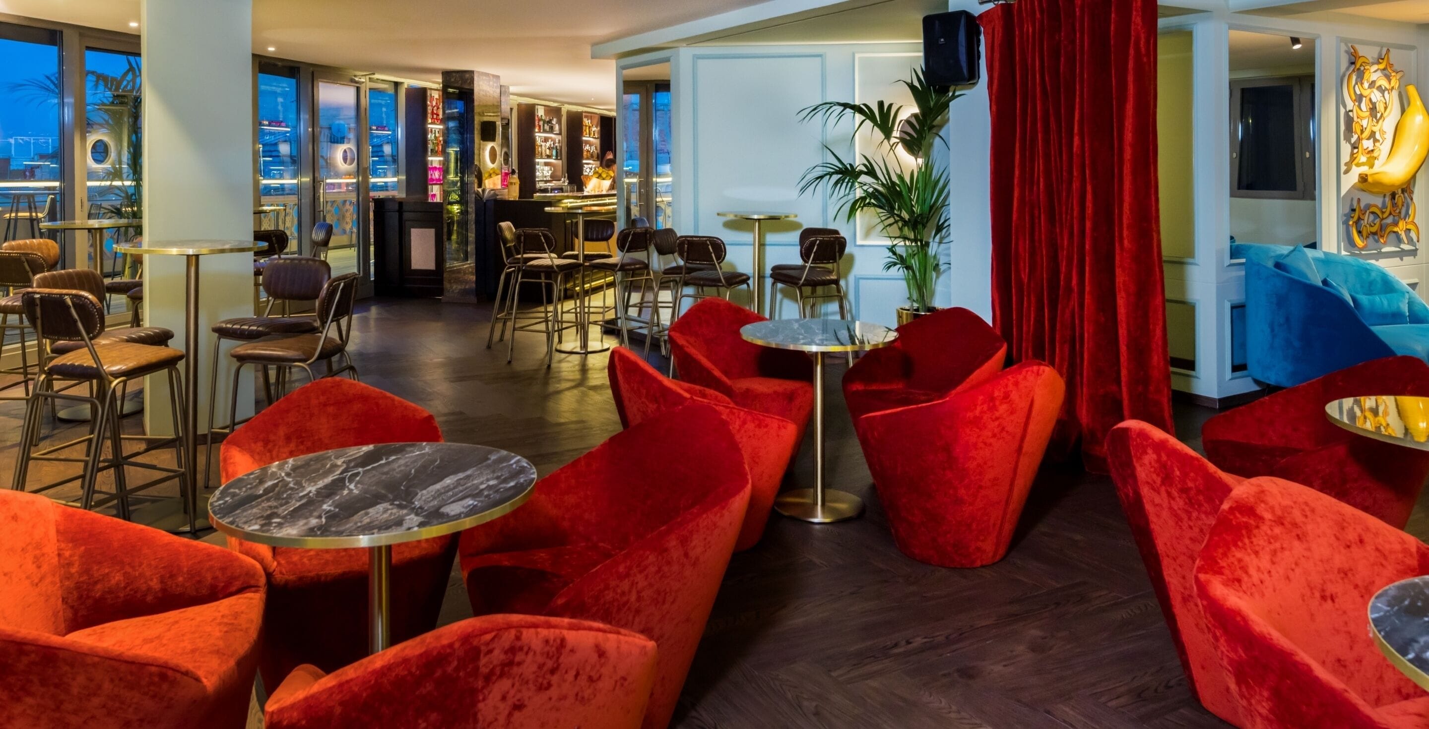 A bar area with red chairs and tables, a bar counter with shelves of bottles, and a large potted plant.