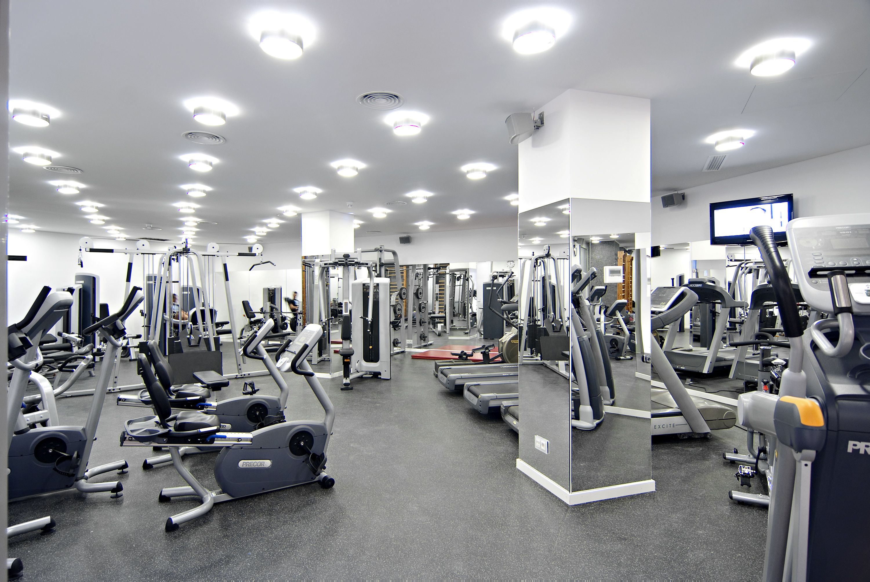 A gym with various exercise machines, mirrors, and a television on the wall.