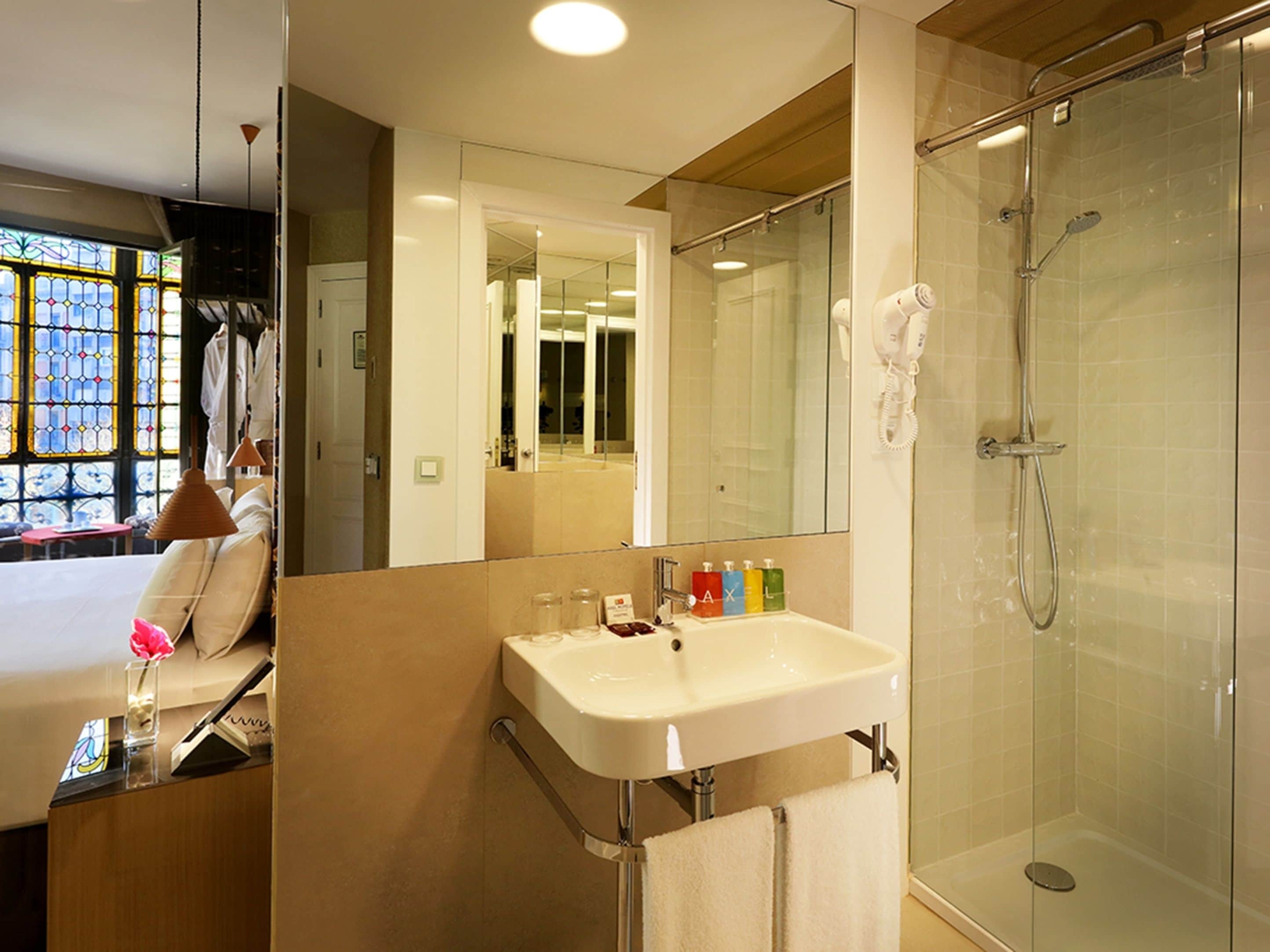 A modern bathroom with a glass shower enclosure, a white sink with a mirror above it, and a window with stained glass.