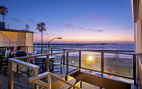 A seaside restaurant with outdoor seating overlooking the ocean, a pier in the background, and a clear sky.