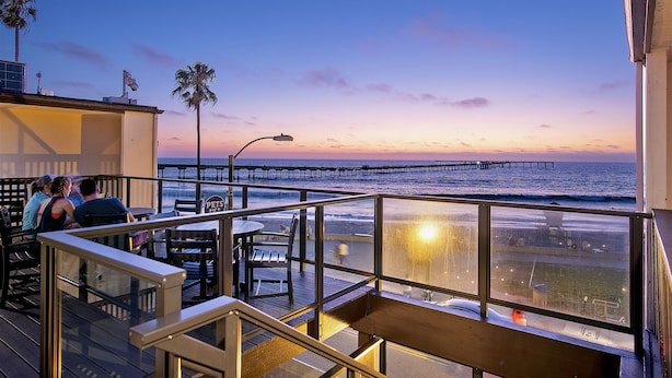 A seaside restaurant with outdoor seating overlooking the ocean, a pier in the background, and a clear sky. - Ocean Beach Hotel (San Diego)