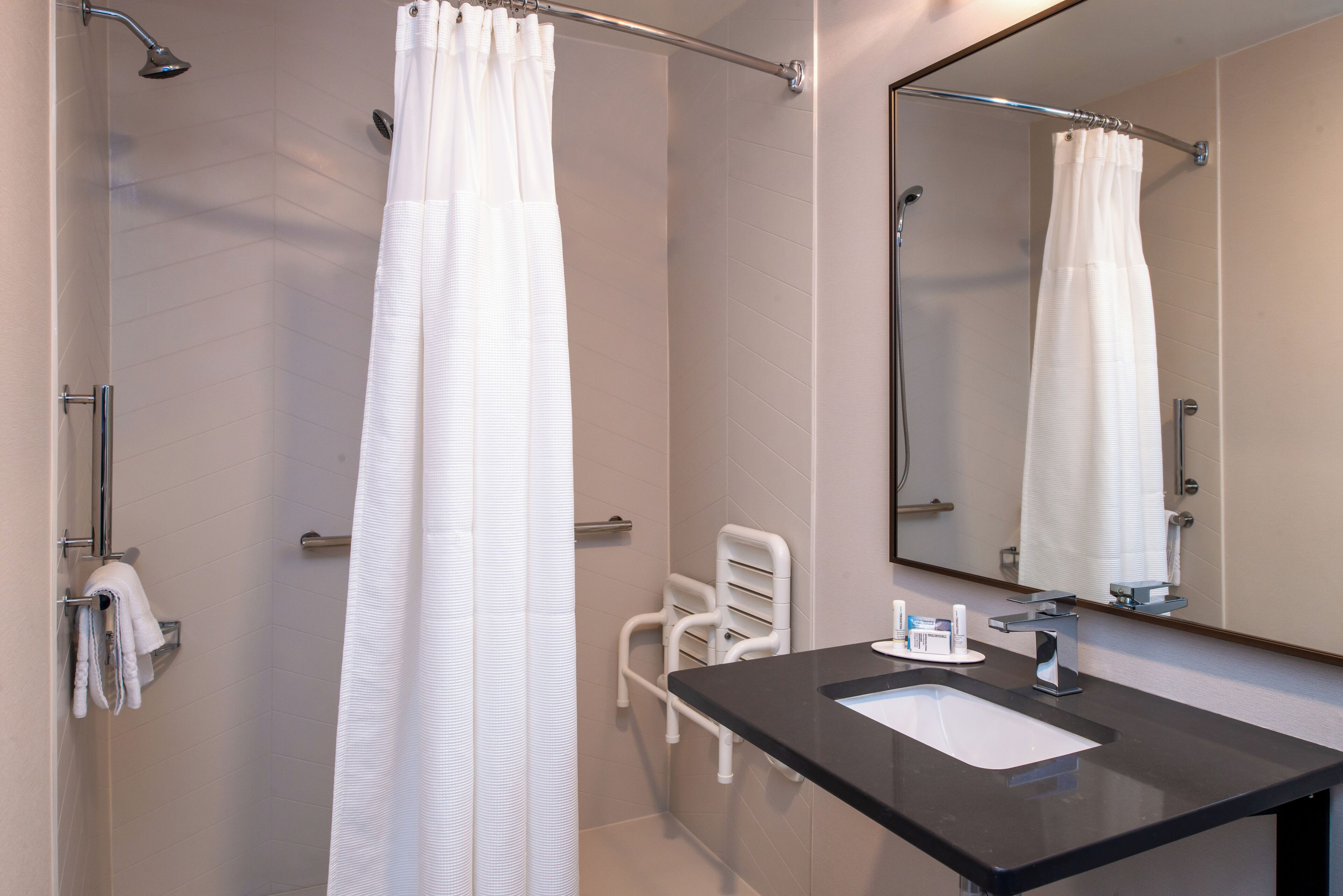 A modern bathroom with a shower, a white towel rack, a white chair, a black countertop, and a mirror.