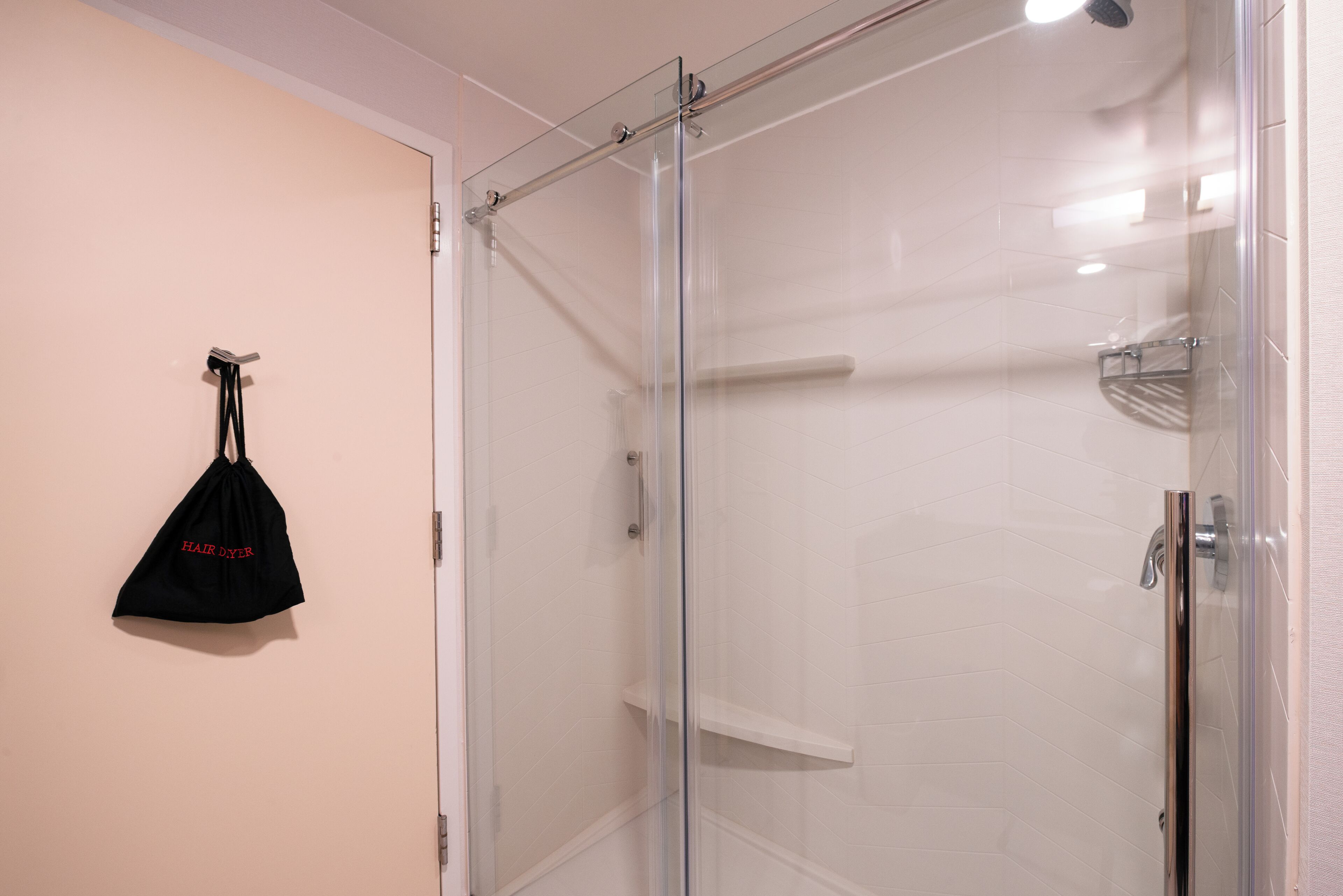 A glass-enclosed shower with a white tiled wall and a black bag hanging on the door.