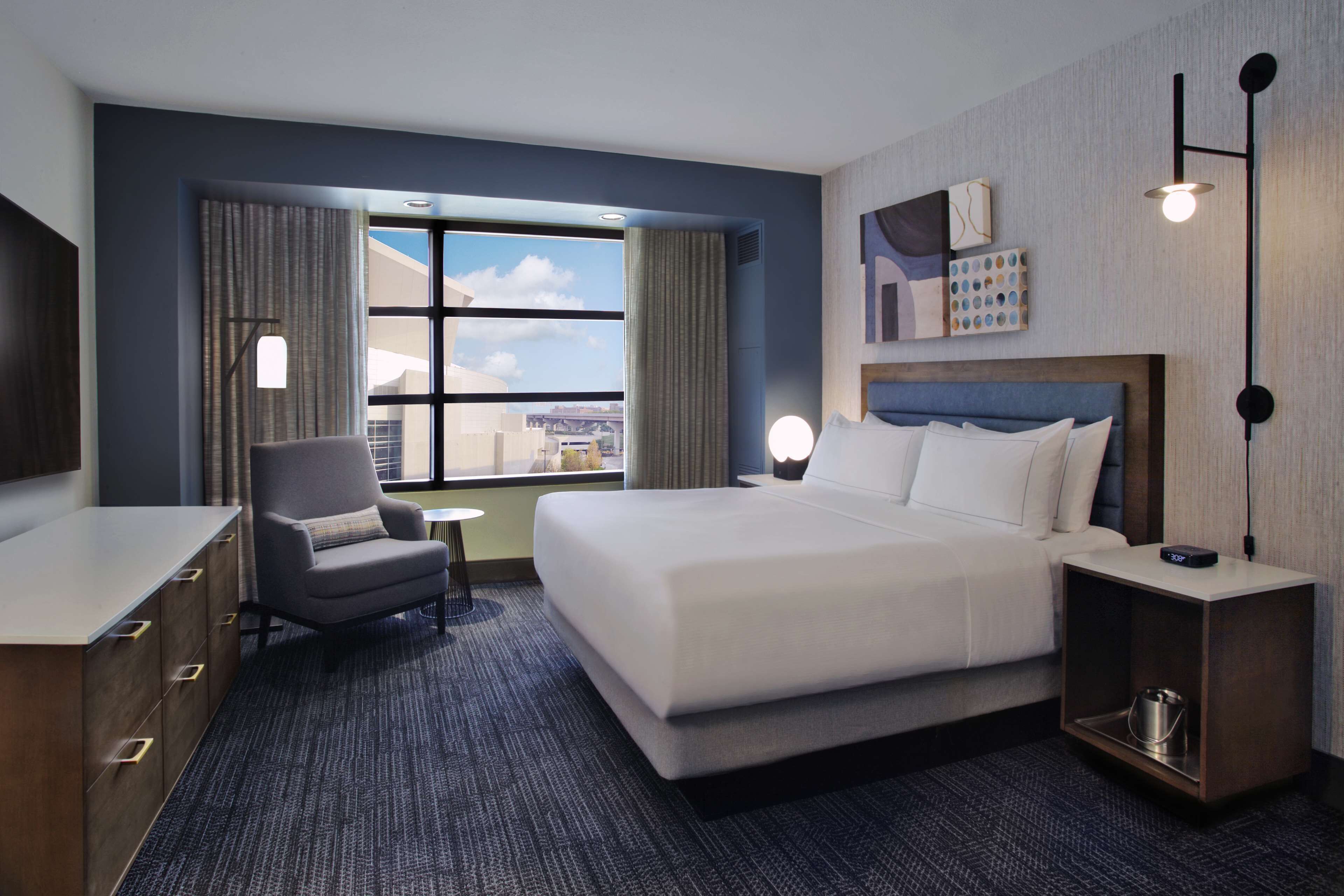 A hotel room with a large bed, a desk, a chair, a small table, a nightstand, and a view of the city through the window.