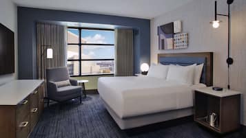 A hotel room with a large bed, a desk, a chair, a small table, a nightstand, and a view of the city through the window.