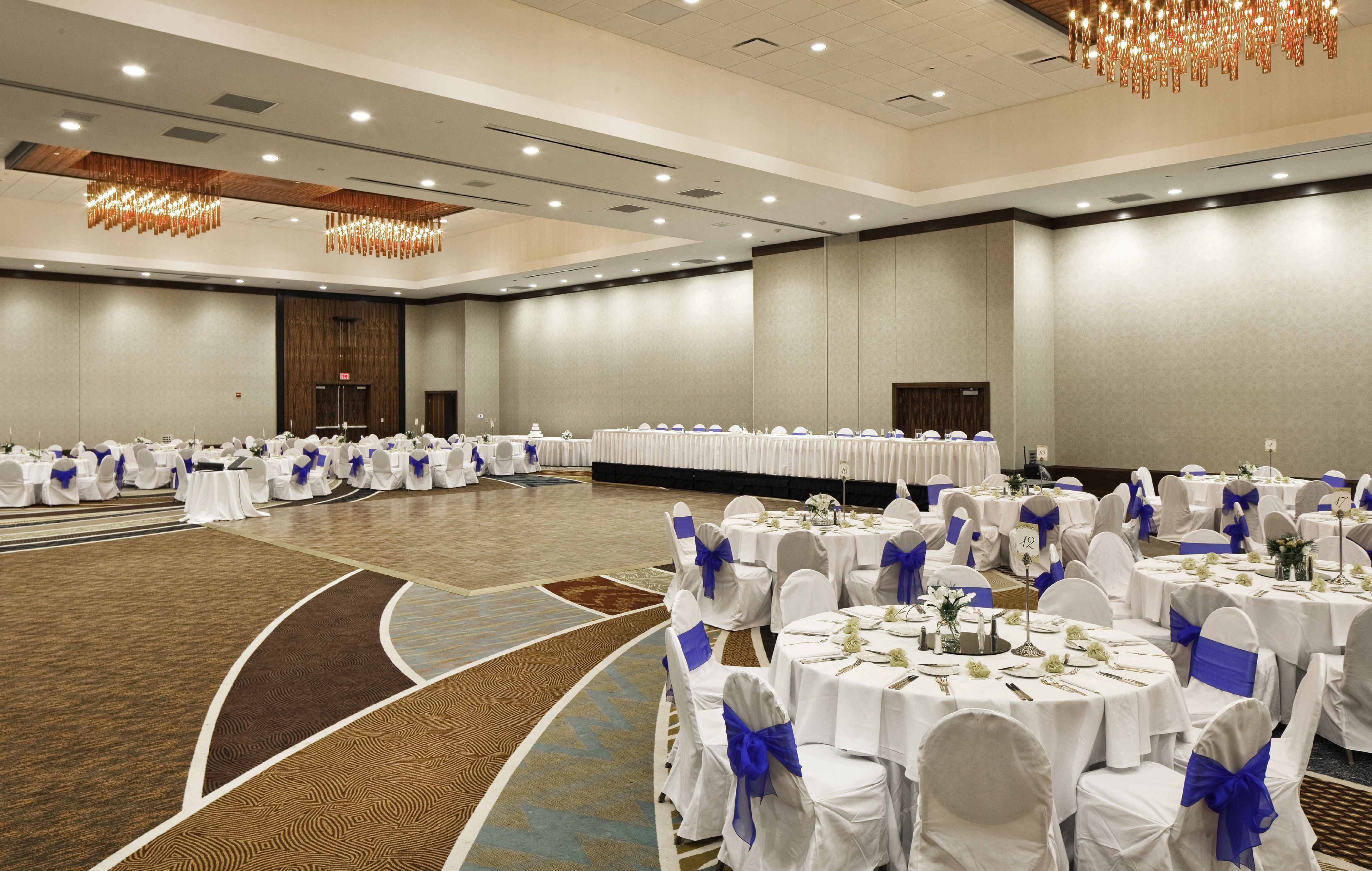 A large, well-lit banquet hall with round tables set for events, featuring white tablecloths and blue sashes.