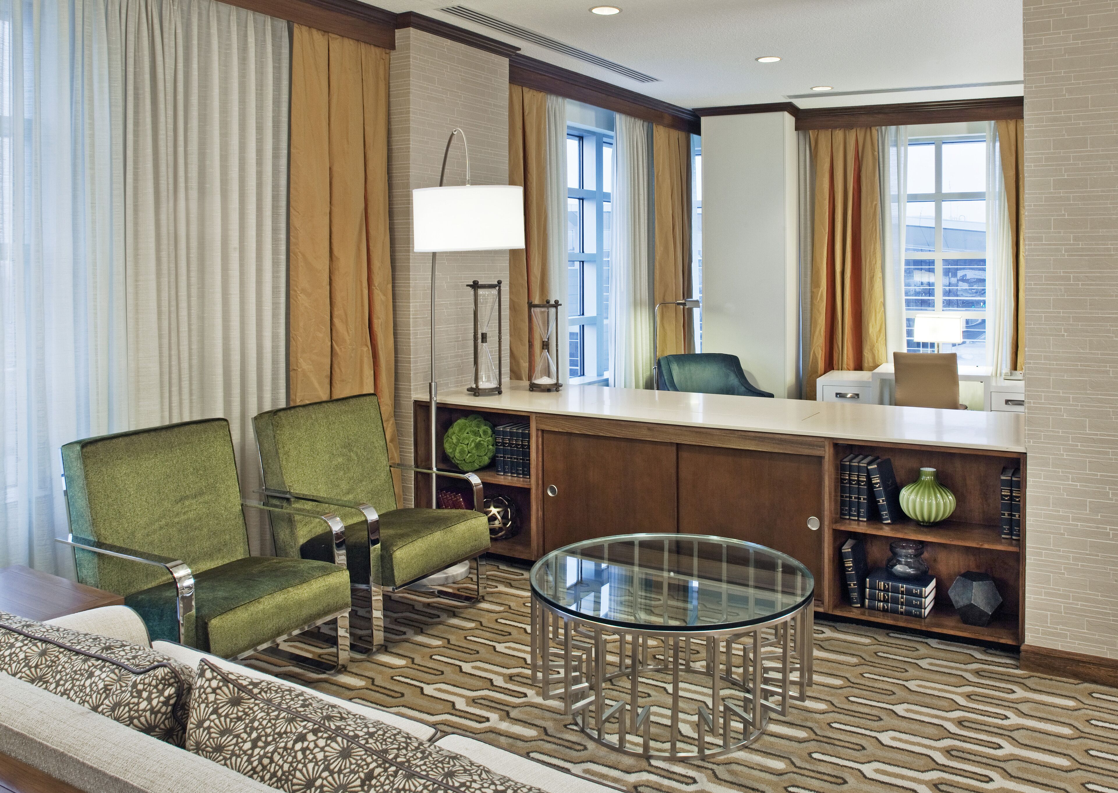 A hotel lobby with a green armchair, a glass coffee table, a wooden desk, and a bookshelf with books and decorative items.