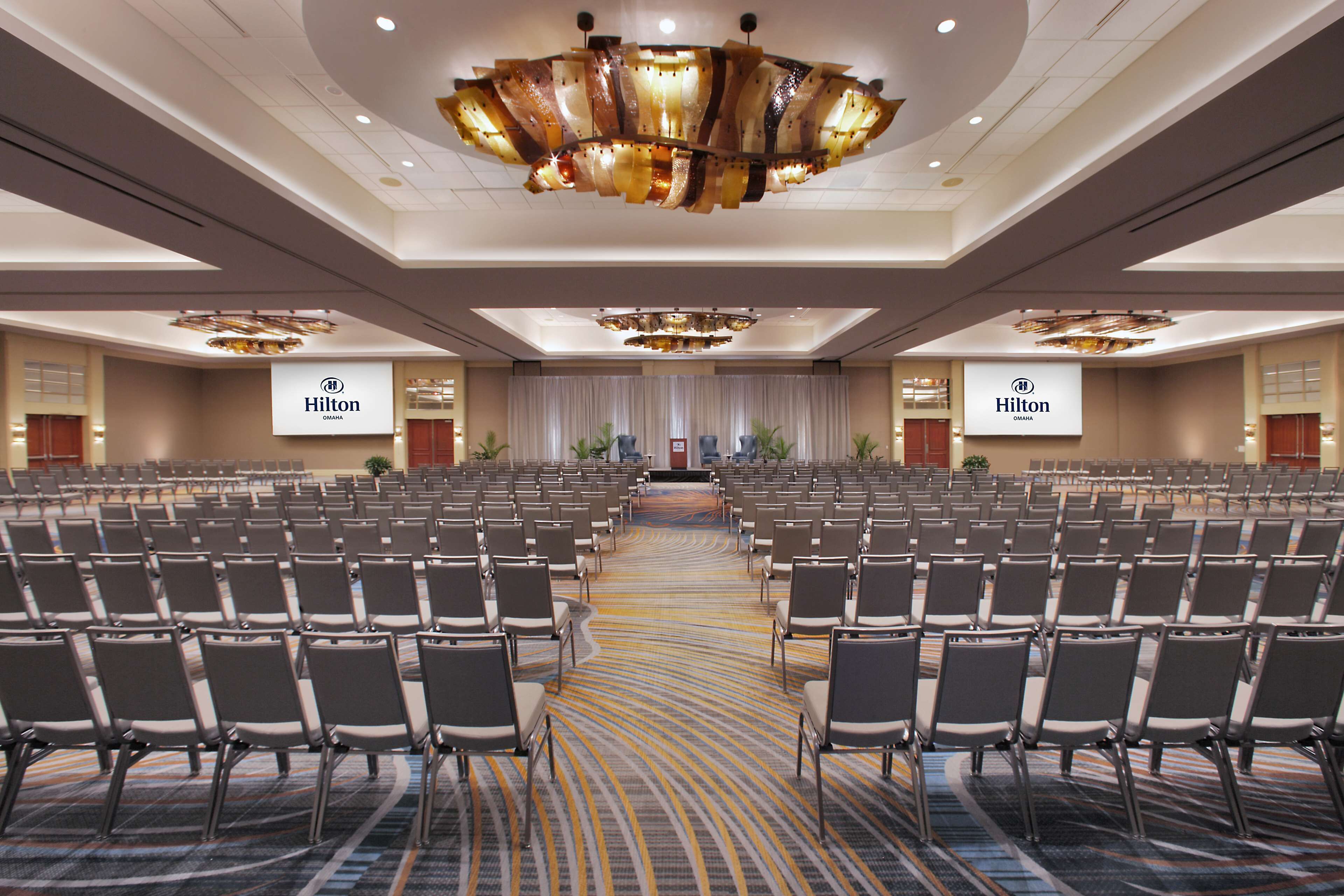 Een grote congreszaal met rijen stoelen, een podium aan de voorzijde, en twee Hilton borden aan de muren.
