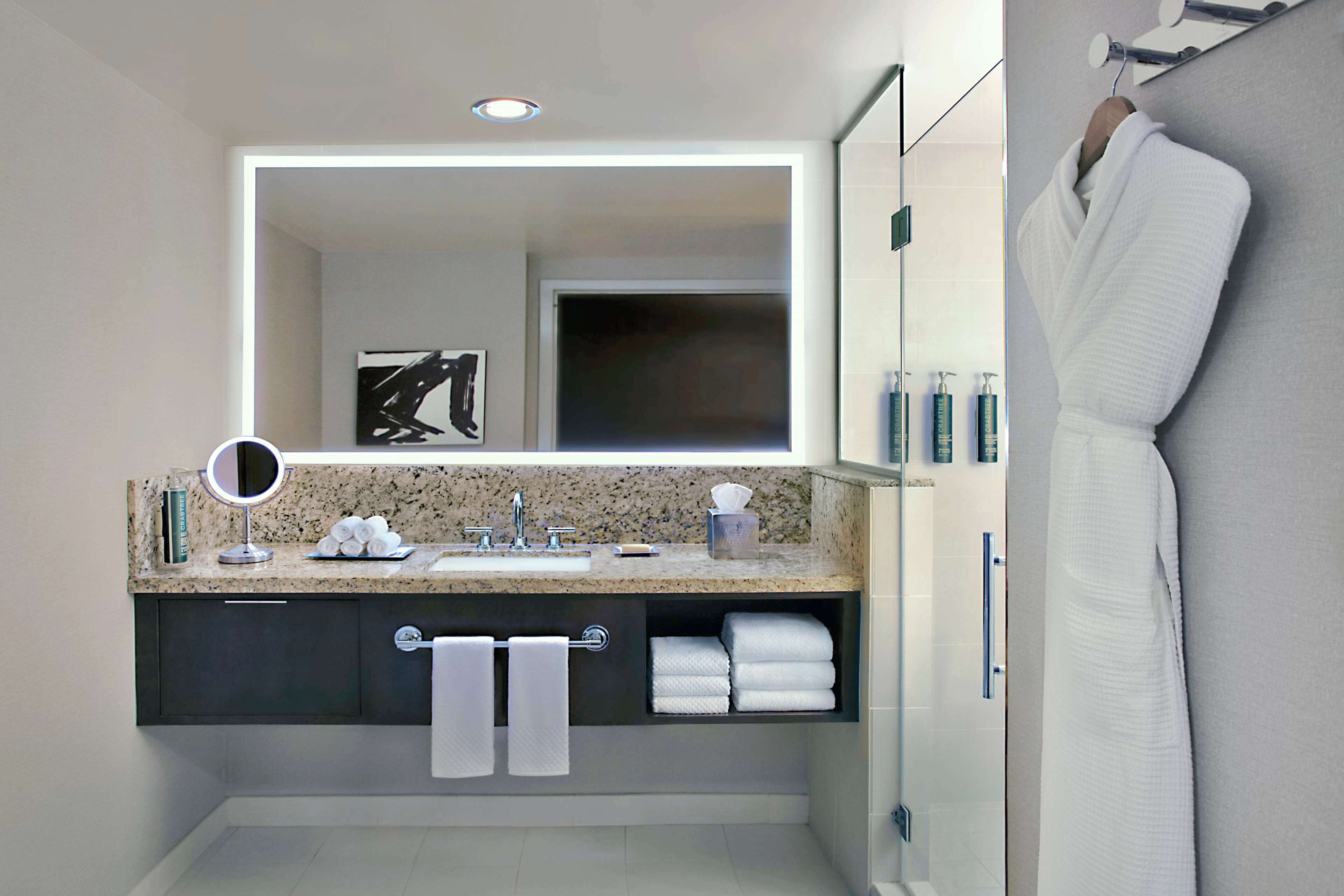 A modern bathroom with a granite countertop, a large mirror, a towel rack, and a glass shower enclosure.
