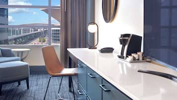 Una habitación de hotel moderna con un ventanal grande, un escritorio, una silla, un espejo y una cafetera.