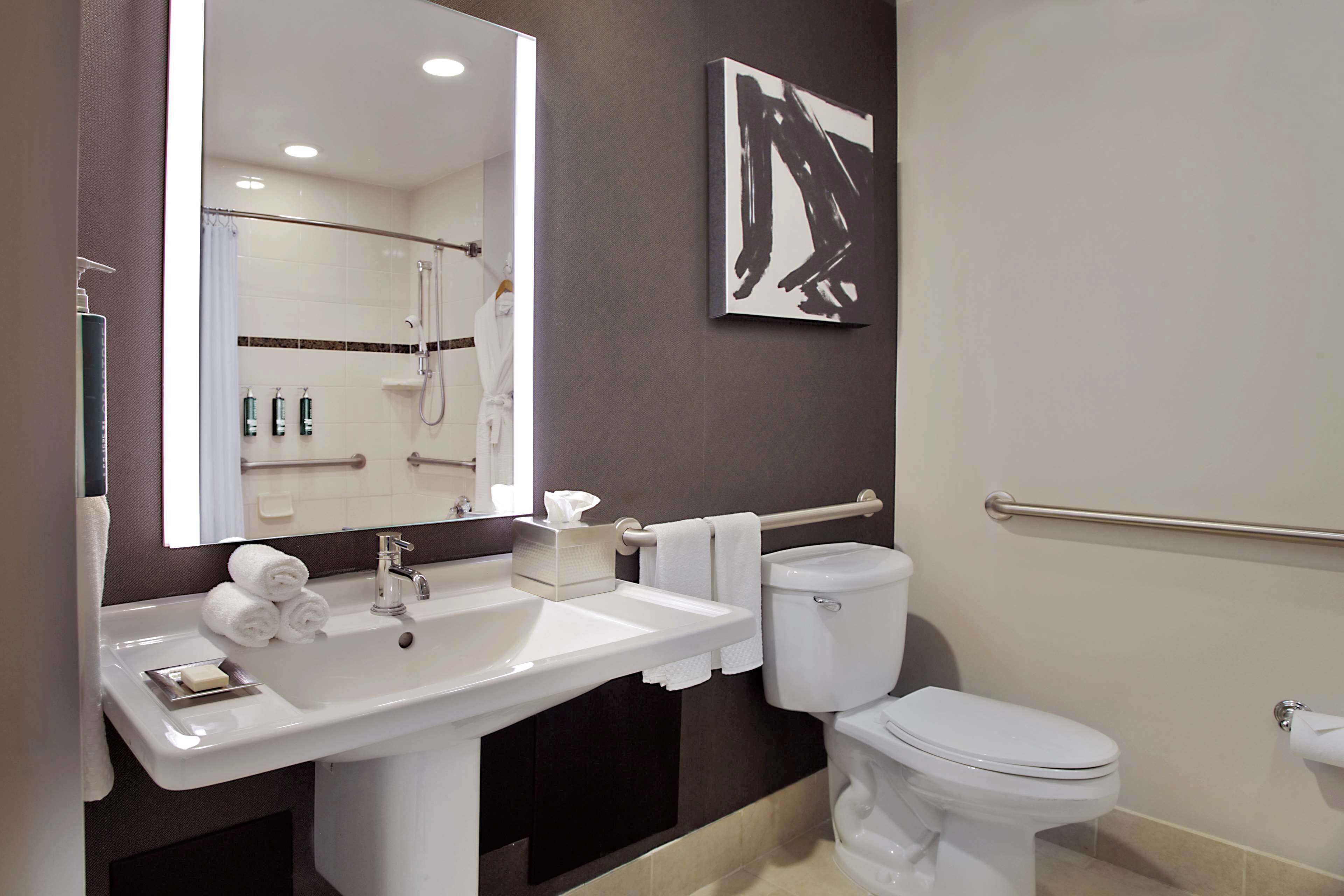A bathroom with a white sink, mirror, and toilet. There is a towel rack with towels, a hand soap dispenser, and a hand dryer. A handrail is mounted on the wall next to the toilet.