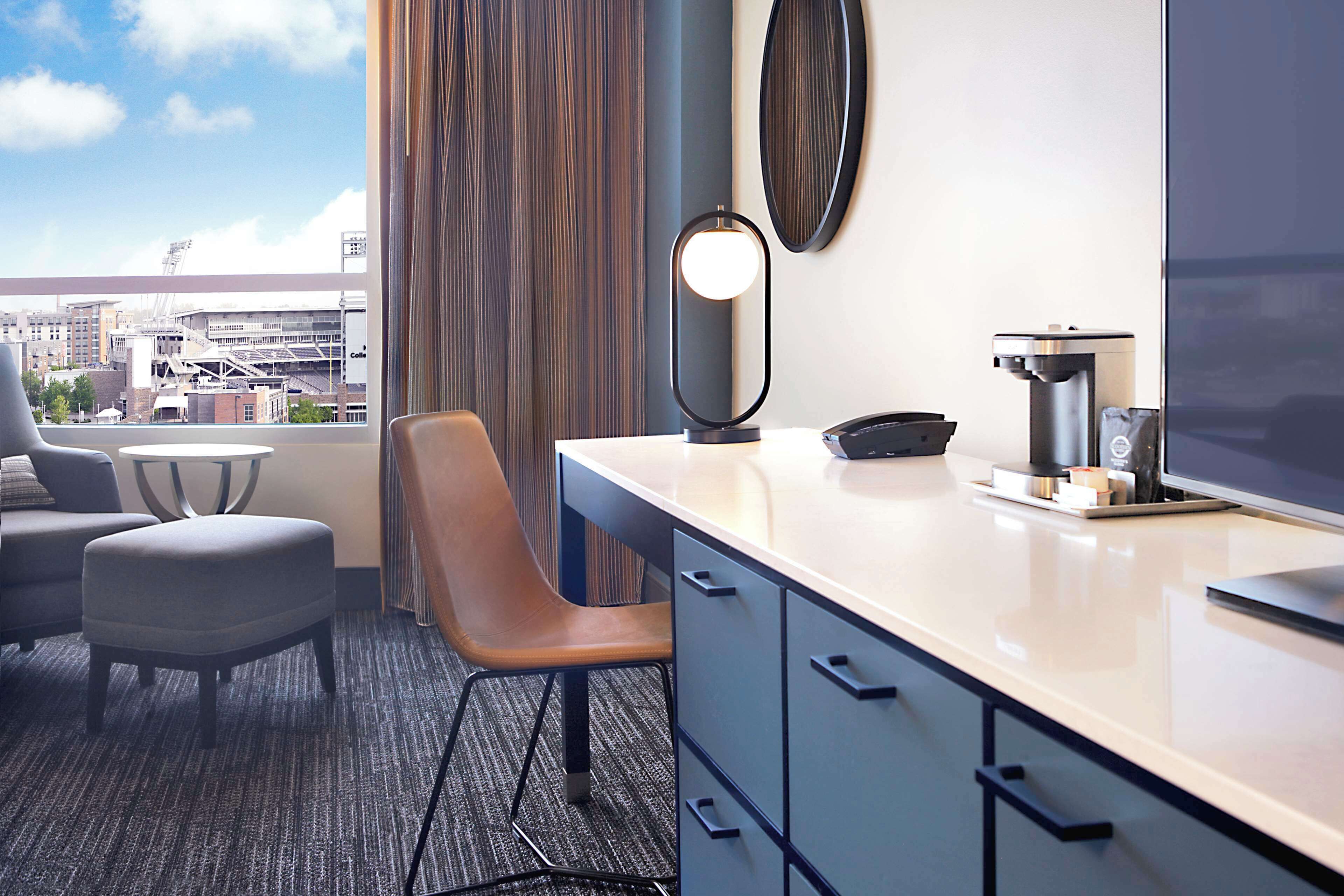 Habitación de hotel con escritorio, silla y vistas a la ciudad a través de amplias ventanas.