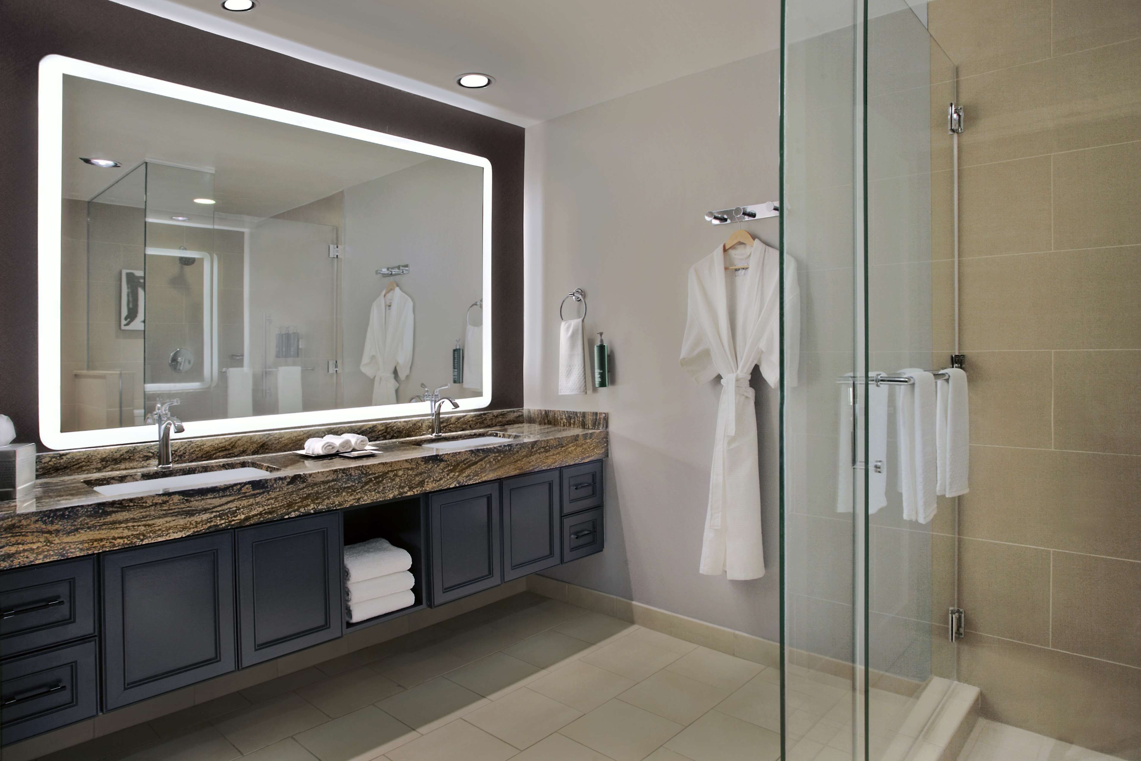A modern bathroom with a large mirror, granite countertop, dark cabinets, and a glass shower enclosure.