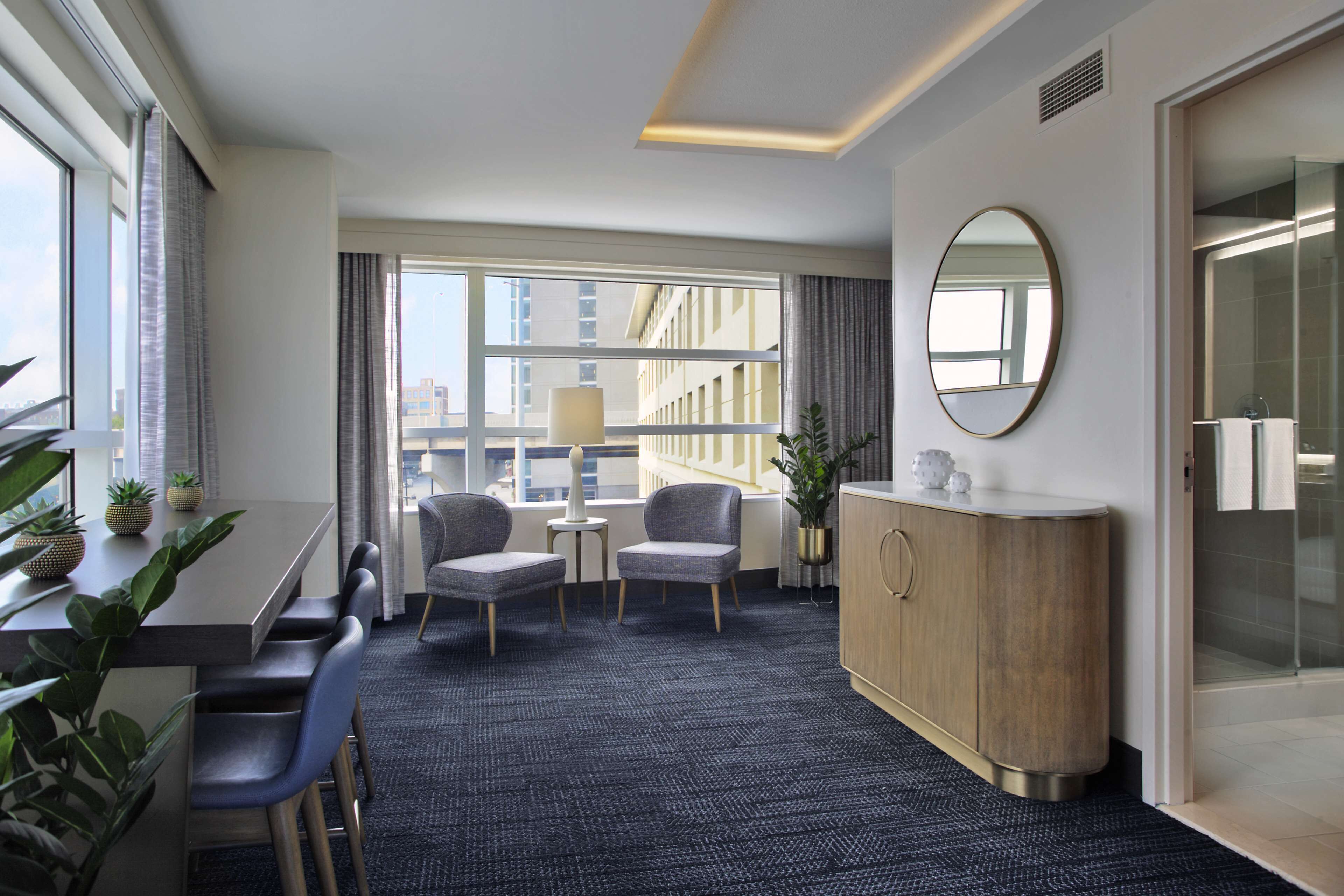A hotel room with a dining area, a large window with curtains, a round mirror, and a wooden cabinet.
