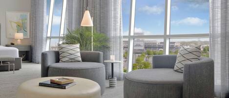 A modern hotel room with a large window, grey upholstered seating, a small table with a lamp, and a framed book on a ottoman.
