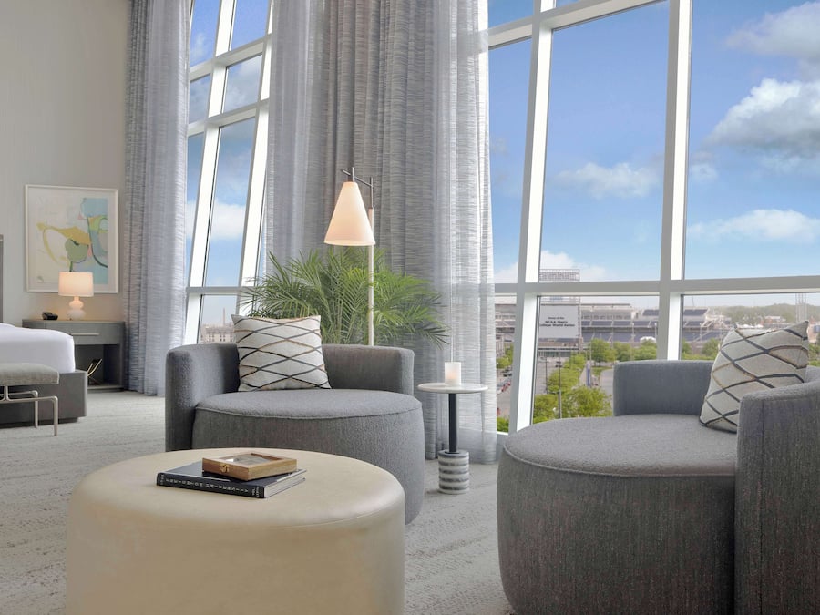 Habitació d'hotel moderna amb una finestra gran, seients tapissats de gris, una taula petita amb una làmpada i un llibre emmarcat sobre un otoman.