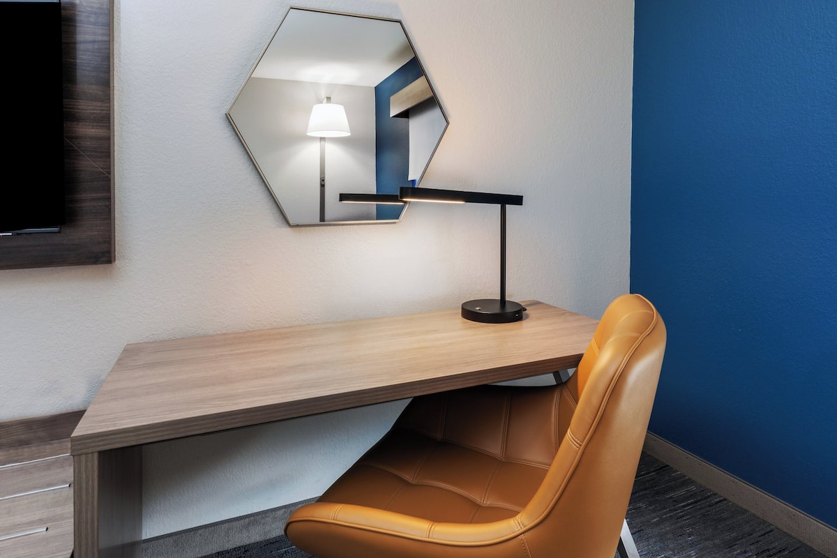 A hotel room with a wooden desk, a leather chair, a wall-mounted mirror, and a TV.
