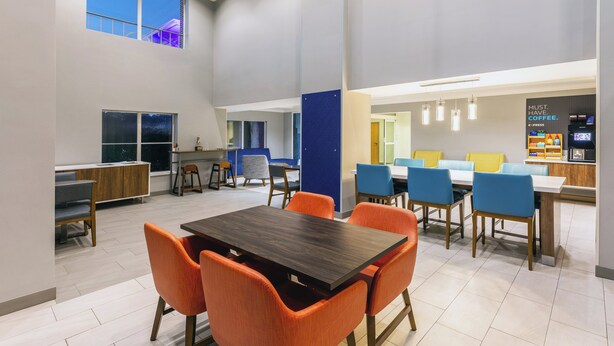 Un espace repas moderne avec une table en bois foncé et des chaises orange, un séparateur bleu et un coin café agrémenté d'une pancarte indiquant : « UN CAFÉ, C'EST INDISPENSABLE ».