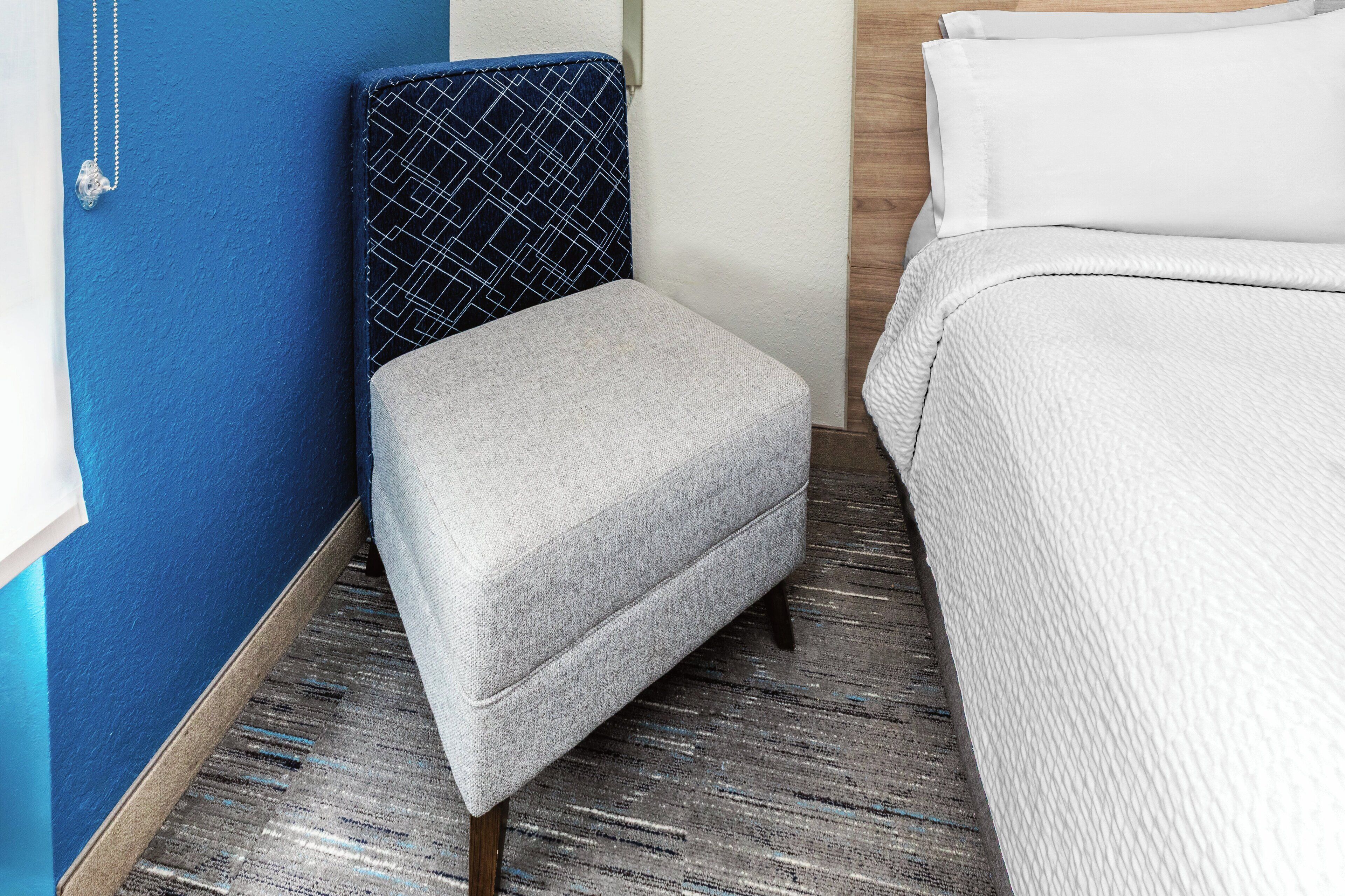 A hotel room with a grey ottoman, a patterned headboard, a white bed, and a blue wall.