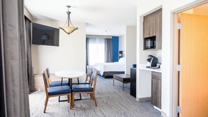 A hotel room with a bed, a desk with a computer, a dining table with chairs, a TV mounted on the wall, and a kitchenette.