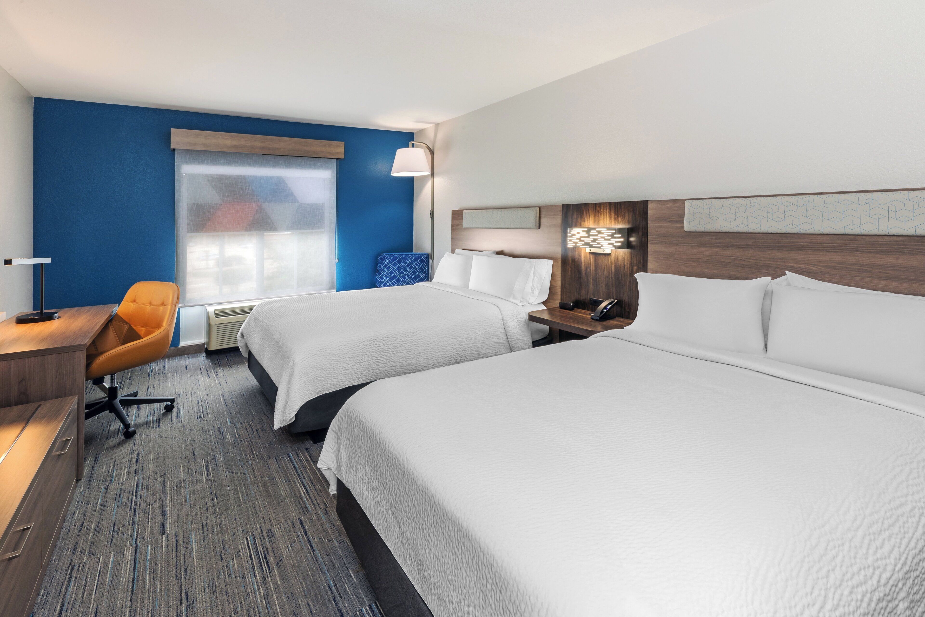 A hotel room with two beds, a desk, a chair, a lamp, and a window.
