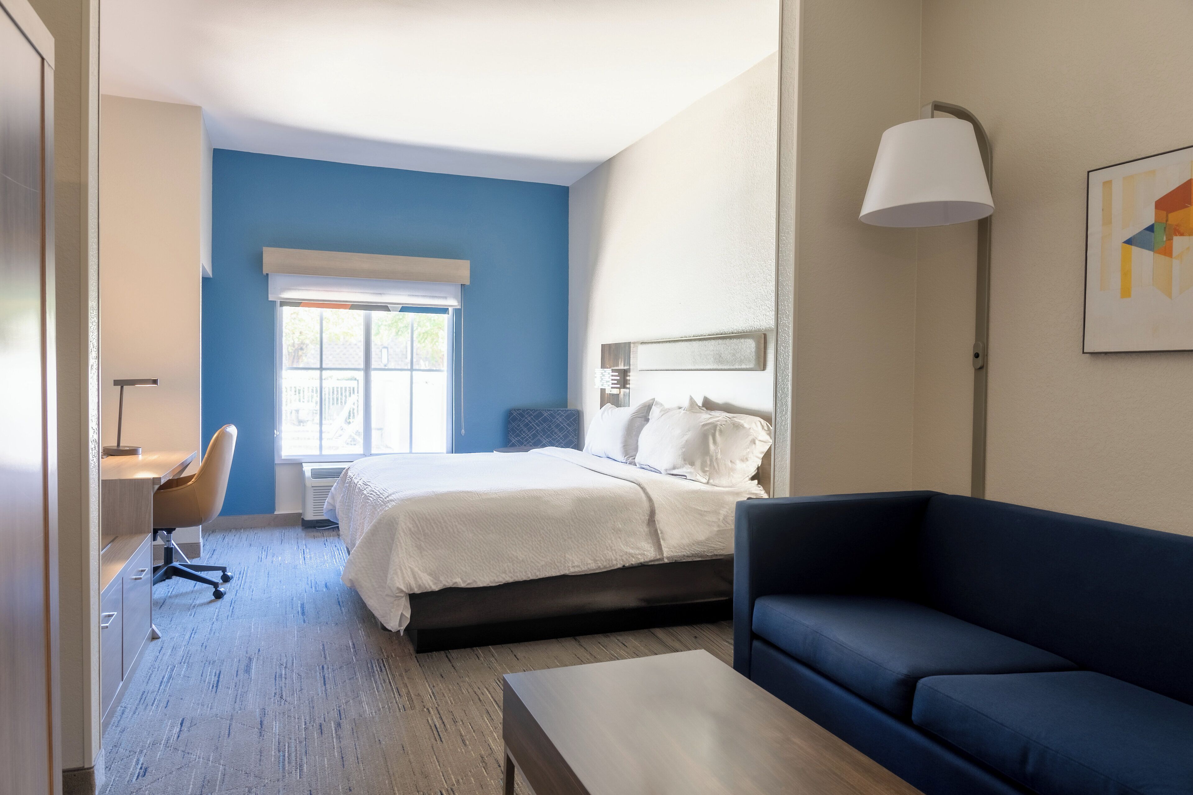 A modern hotel room with a bed, a desk, a chair, a sofa, and a lamp.