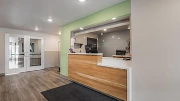 Un espace d’accueil avec un comptoir de réception en bois, une porte vitrée donnant sur un couloir et, au fond, un coin cuisine.