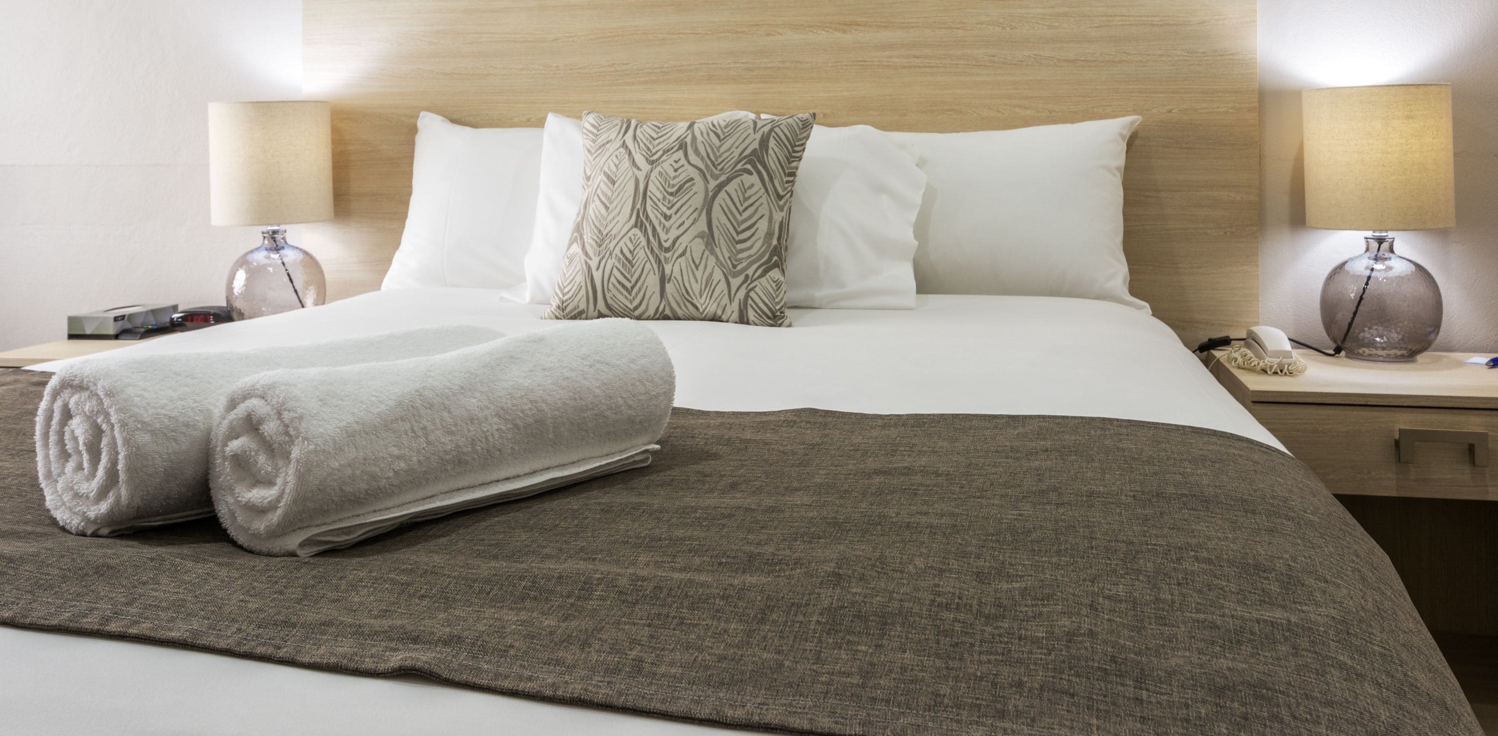 Un lit bien fait, recouvert d’une couverture marron, avec deux serviettes blanches et un oreiller à motifs. On y trouve deux lampes de chevet et un téléphone sur la table de nuit.