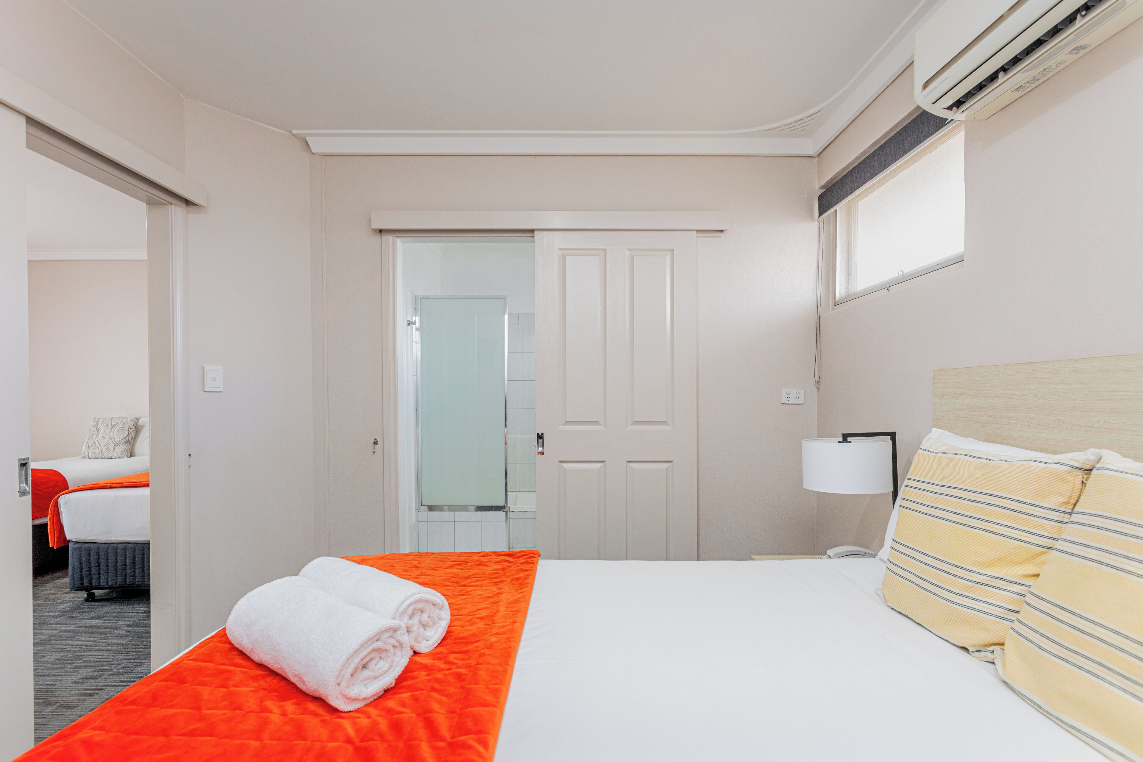 A hotel room with a bed, a nightstand, a lamp, and a door leading to another room.