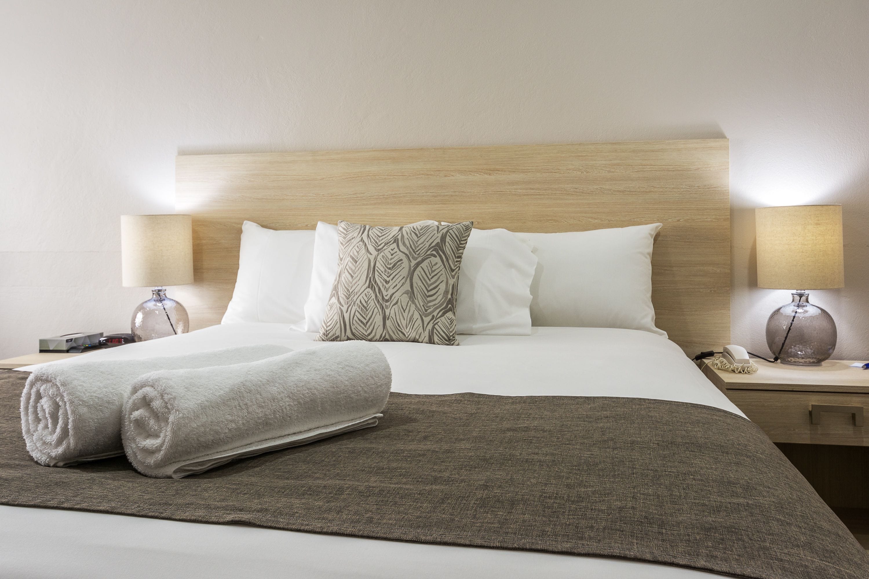 Un lit bien fait, avec des oreillers blancs et à motifs, une serviette blanche roulée et un plaid marron. Il y a deux tables de chevet, chacune avec une lampe, et un vase sur l’une d’elles.