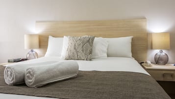 Un lit bien fait, avec des oreillers blancs et à motifs, une serviette blanche roulée et un plaid marron. Il y a deux tables de chevet, chacune avec une lampe, et un vase sur l’une d’elles.
