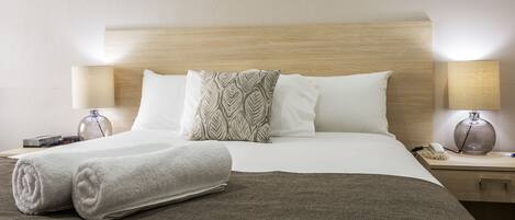 A neatly made bed with white and patterned pillows, a rolled white towel, and a brown throw blanket. There are two bedside tables with lamps and a vase on one.