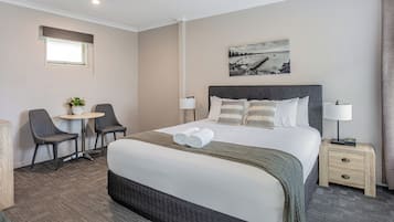 A hotel room with a large bed, two chairs, a small table with a plant, a nightstand with a lamp, and a painting on the wall.