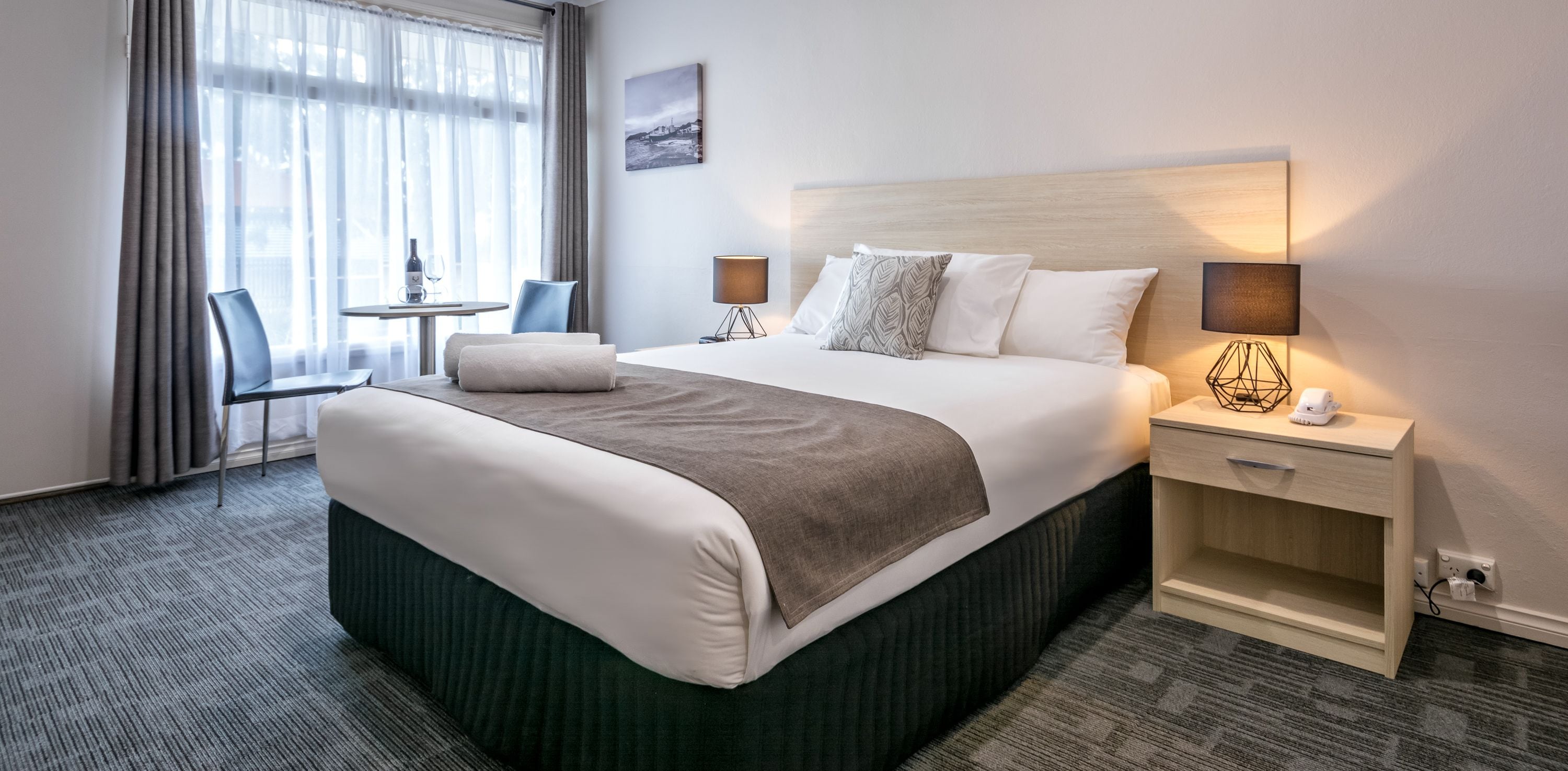 A hotel room with a large bed, two bedside tables with lamps, a small table with a chair, and a window with curtains.