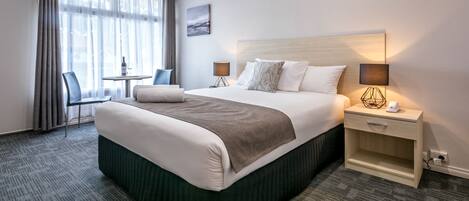 Habitación de hotel con una cama grande, dos mesitas de noche con lámparas, una pequeña mesa con una silla y una ventana con cortinas.