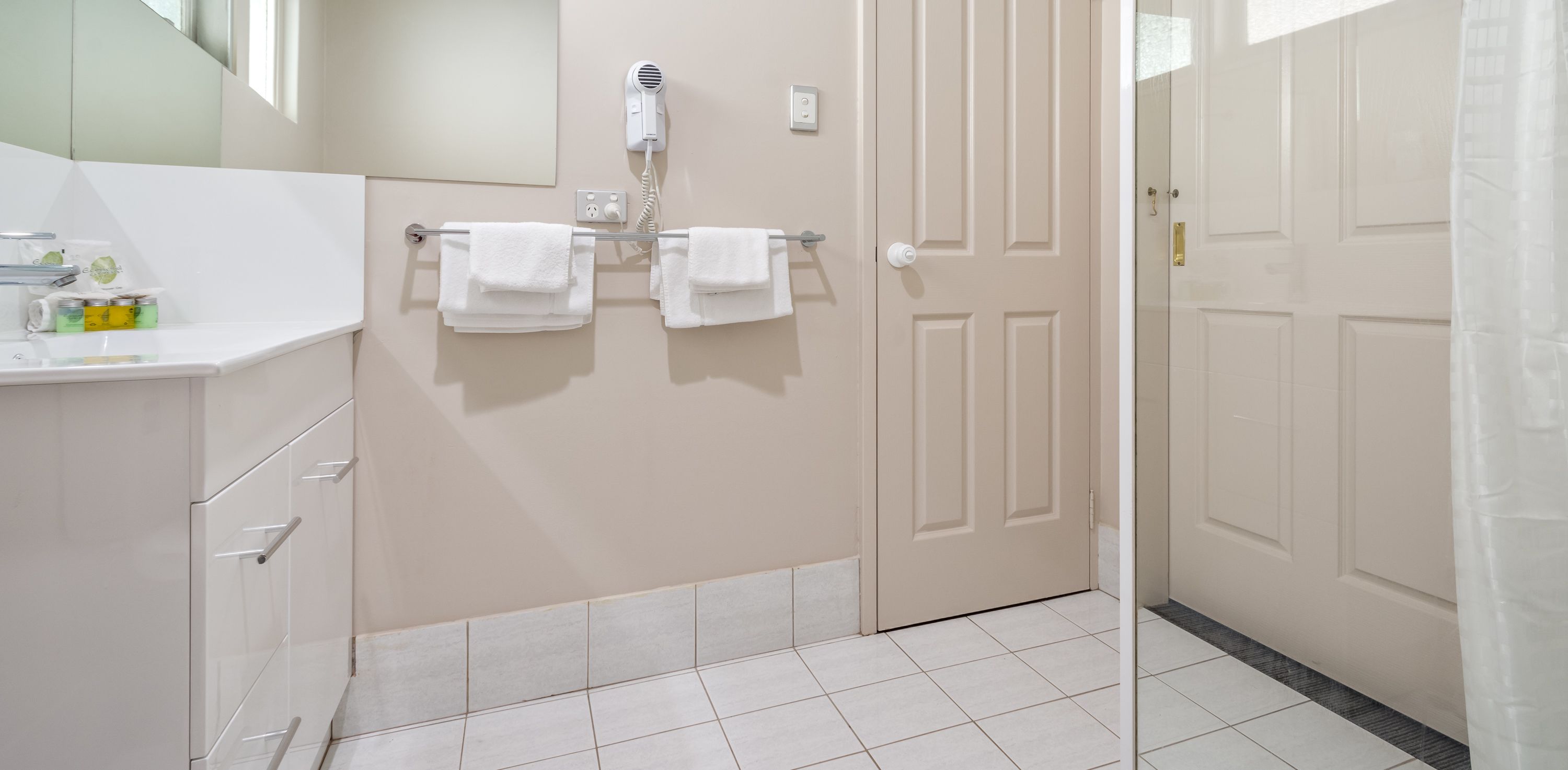 Un bagno con un lavandino bianco, due asciugamani appesi a un portasciugamani, un asciugacapelli montato sulla parete e una porta con maniglia dorata.