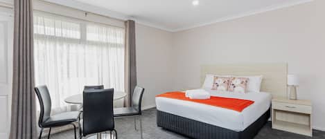 Una habitación de hotel con cama, mesita de noche, silla, una pequeña mesa y ventana con cortinas.