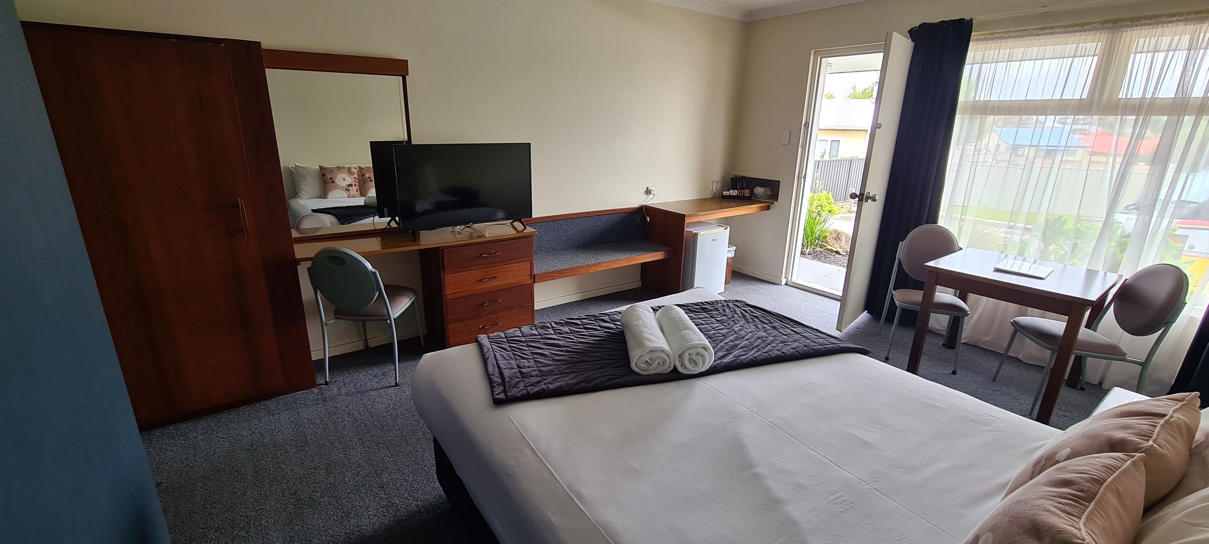 A hotel room with a bed, a desk with a television, a chair, a small table, and a view through a door.