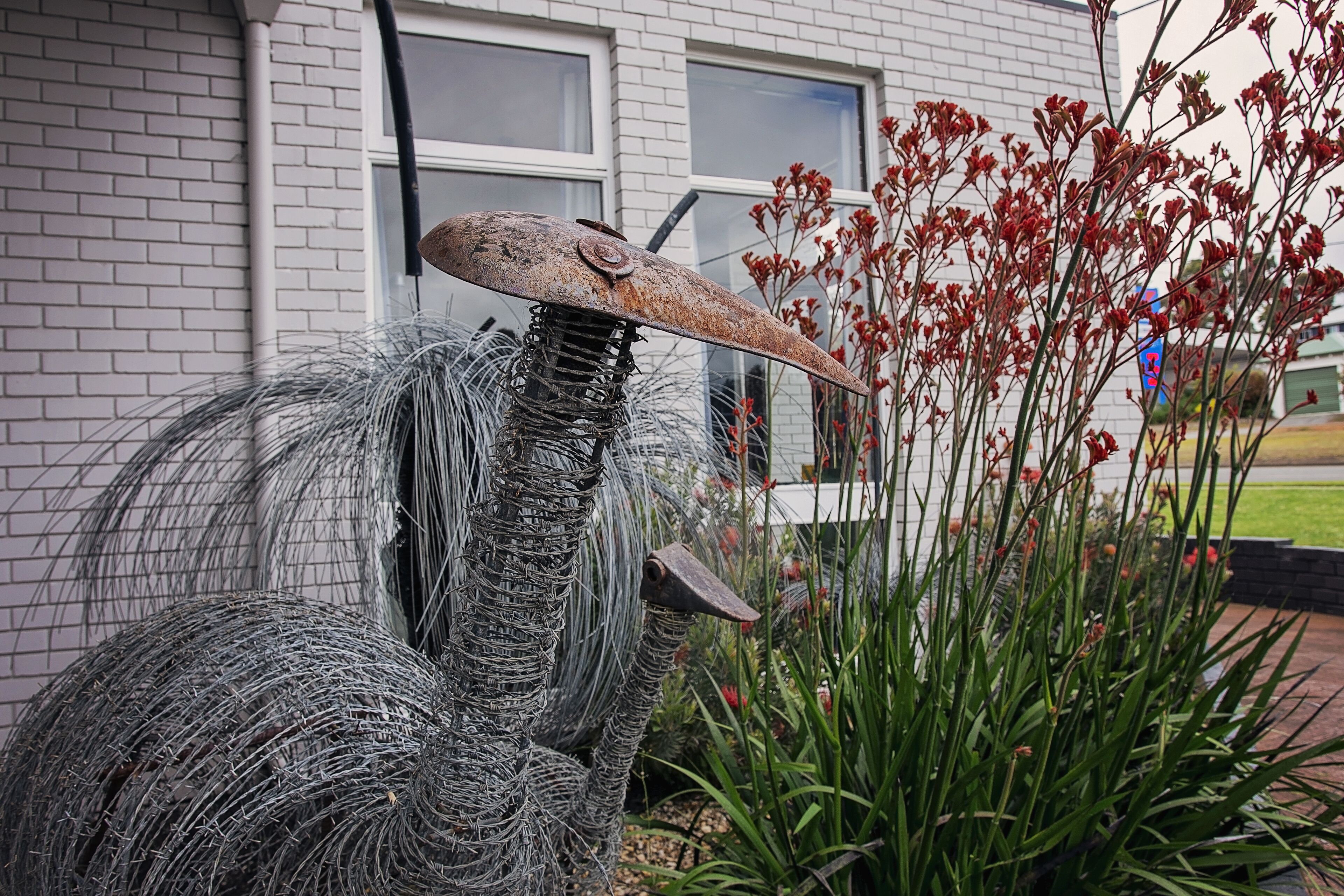 A sculpture of a bird with a large beak made of wire mesh, set against a brick building with windows and red flowers in the background.
