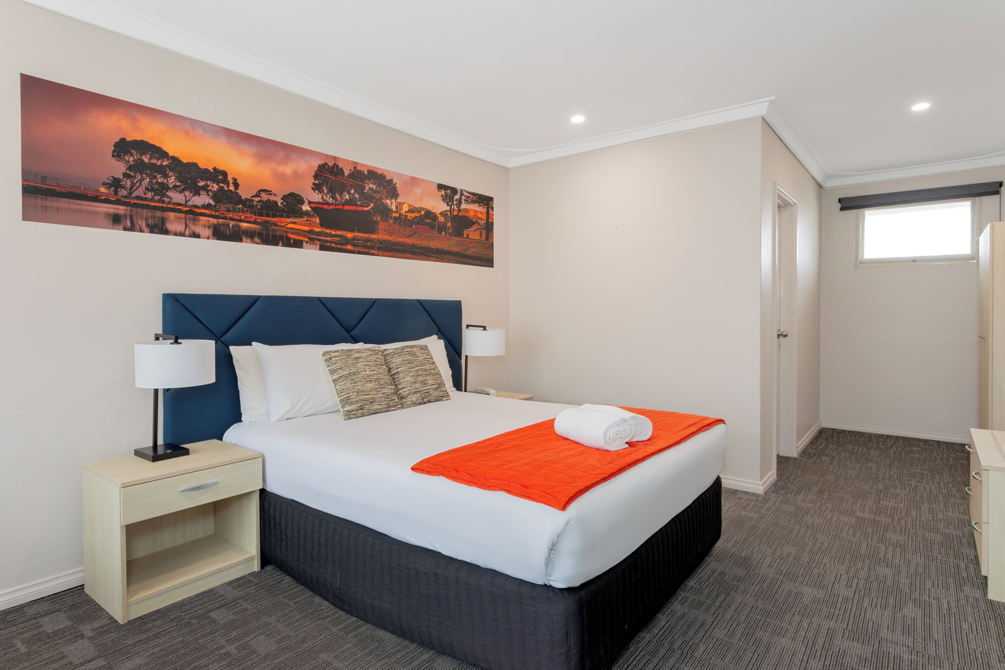 Una cama bien hecha con un cabecero azul, ropa de cama blanca y una manta naranja. Hay una mesita de noche con una lámpara y un estante pequeño. Un cuadro de paisaje grande adorna la pared sobre la cama.